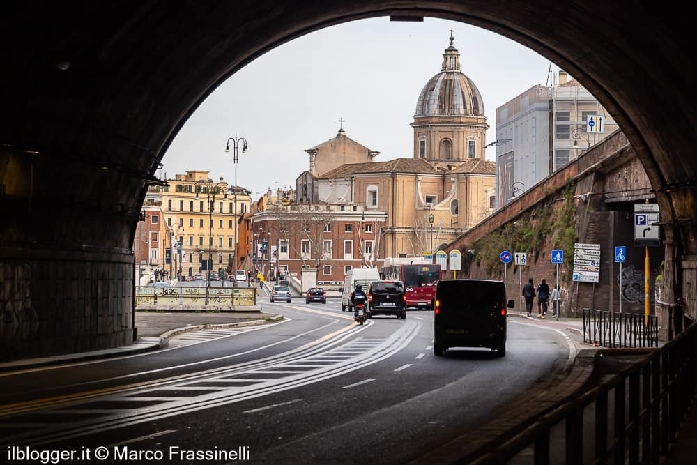 Veduta urbana di Roma incorniciata da un sottopasso, con traffico cittadino in primo piano e la cupola di una chiesa storica sullo sfondo